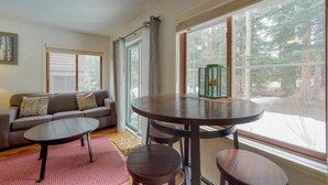 A living room with a sofa, a round table, and a view of a forest through large windows. - Breckenridge Ski-in, Hot Tub, Pet-Friendly (Breckenridge)