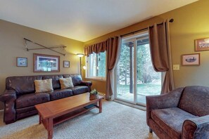 A living room with a brown leather sofa, a wooden coffee table, and a patterned armchair. There are framed pictures on the wall and a large window with curtains.