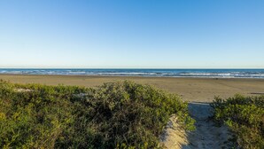 A beach with a clear sky, sandy shore, and dense vegetation in the foreground. - 1st row 4 bedroom 3 bath - Best views! (Galveston)