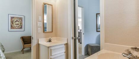 A bathroom with a white vanity, a sink, and a mirror. There is a framed picture on the wall and a chair in the corner.