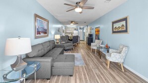 A spacious living area with a grey sectional sofa, a glass coffee table, and a patterned armchair. There are ceiling fans, a dining table with chairs, and framed artwork on the walls.