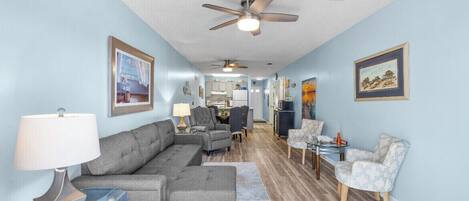 A spacious living area with a grey sectional sofa, a glass coffee table, and a patterned armchair. There are ceiling fans, a dining table with chairs, and framed artwork on the walls.