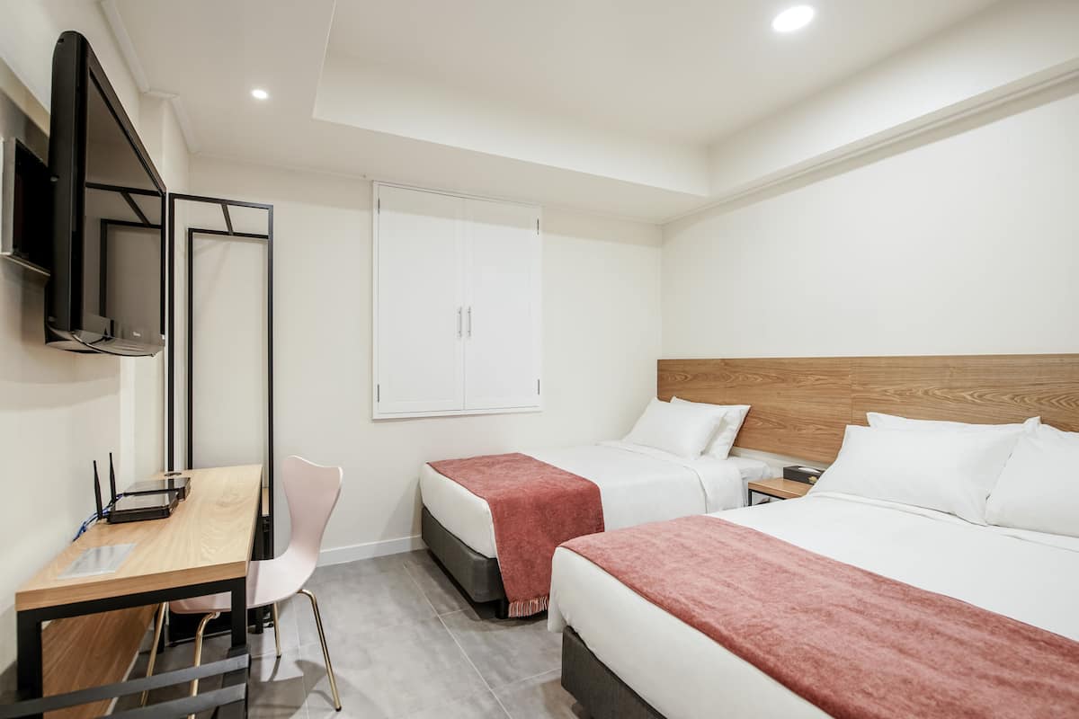 A hotel room with two beds, a desk with a computer, a TV mounted on the wall, and a wooden headboard.