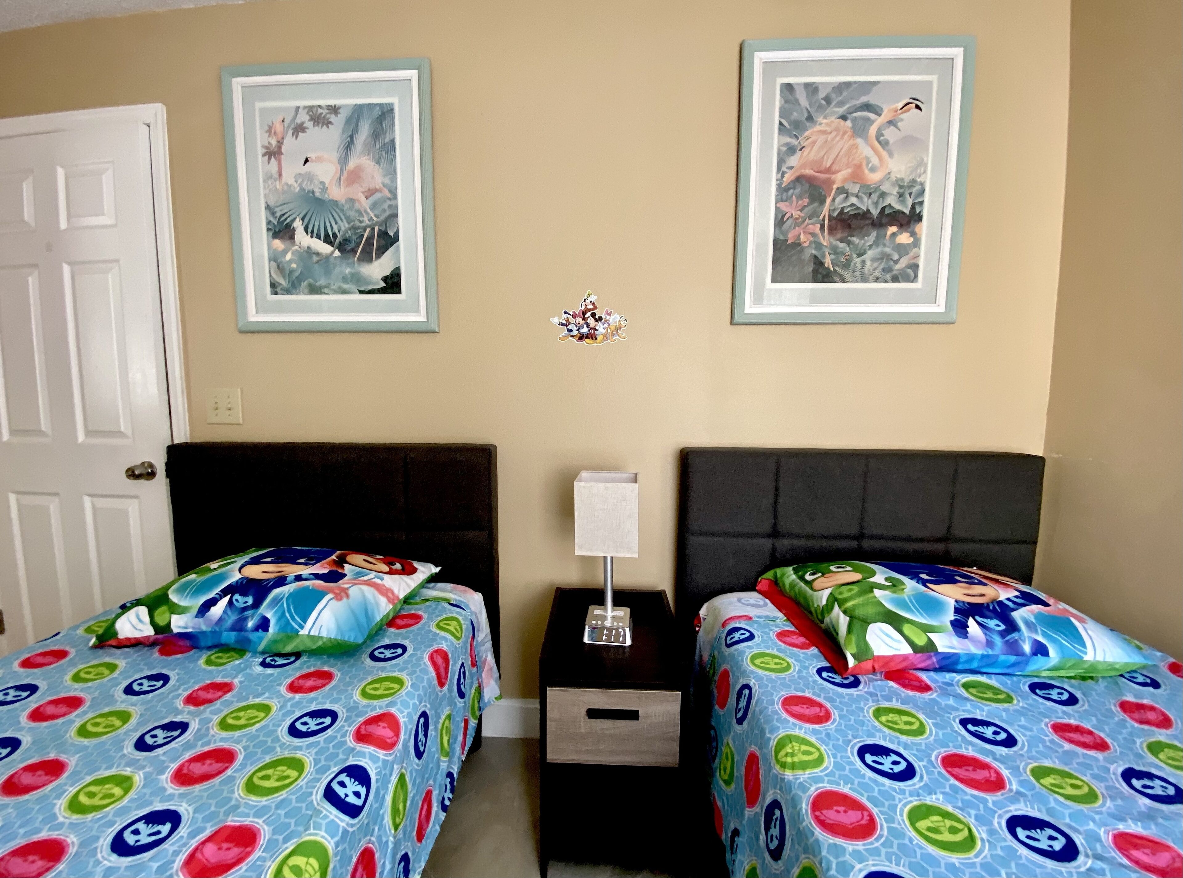 A bedroom with two beds, each with colorful bedding featuring cartoon characters. There are two framed pictures on the wall, one depicting flamingos and the other a cartoon character. A nightstand with a lamp and a small drawer is between the beds.