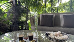 A glass table with two cups of coffee, a decorative bowl, and a wicker chair with cushions in a lush, green outdoor setting. - Poé Côté Lagon Beach Bungalows (Bourail)