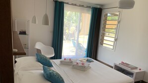 A neatly made bed with blue pillows, a bedside table with a book, and a window with curtains. - Poé Côté Lagon Beach Bungalows (Bourail)