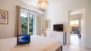 A bedroom with a bed, a laptop, a TV, and a balcony with a view of greenery. - CASA BUGANVILLA 5 in Minia (Argostoli)