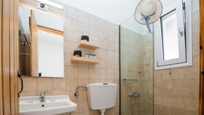 A bathroom with a sink, toilet, and shower. There is a mirror above the sink and a shelf with toiletries. - CASA BUGANVILLA 5 in Minia (Argostoli)