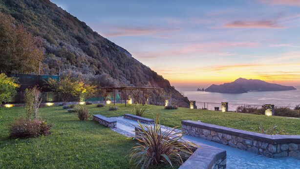 Un giardino curato con una pergola, sedute in pietra e vista sul mare al tramonto.
