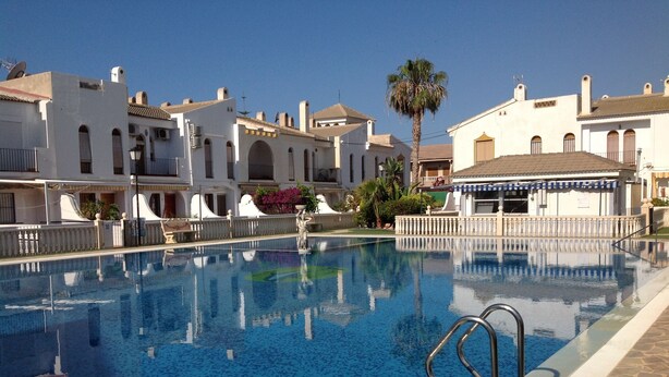 A swimming pool with clear blue water, surrounded by white buildings with arched windows and balconies, and a palm tree in the background. - Delightful family villa in secure gated community with large pool. (Puerto De Mazarrón)