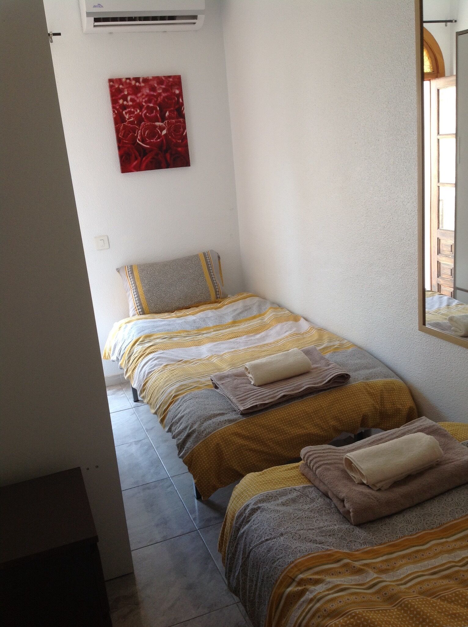 A small, neatly arranged bedroom with two beds, a red artwork on the wall, and a window providing natural light.