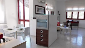 Una cocina compacta con horno y refrigerador integrados, una mesa de comedor con sillas y un sofá en una sala de estar con vistas a través de grandes ventanales.