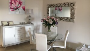 Un coin repas avec une table en verre, des chaises blanches, un buffet blanc, un tableau représentant des fleurs, une lampe et un miroir doté d’un cadre orné.