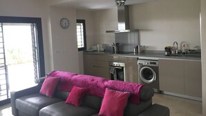 Une cuisine moderne équipée d’un four encastré, d’un four à micro-ondes et d’un lave-linge. Le salon est doté d’un canapé gris agrémenté de coussins roses.