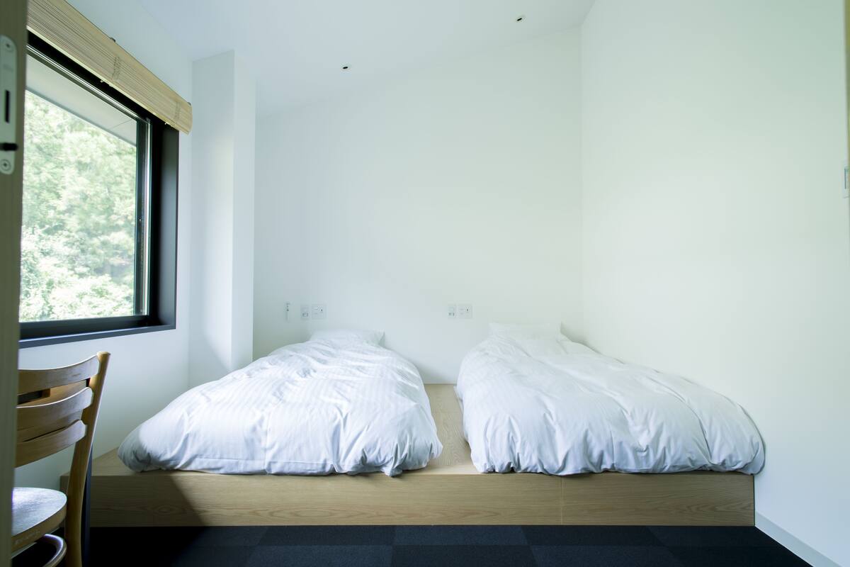 A room with two beds, a wooden chair, and a window with a view of trees.