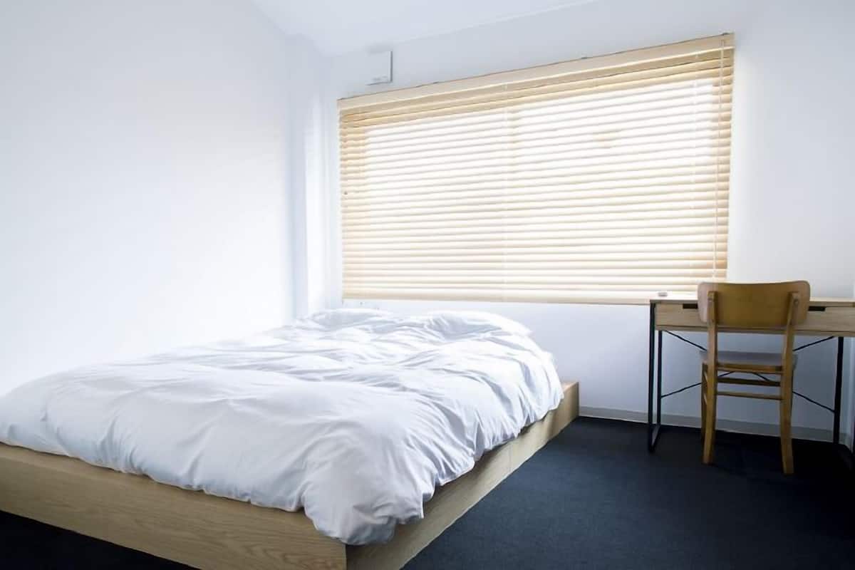 A single bed with a wooden frame, a desk with a chair, and a window with blinds.