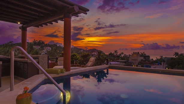 Piscina con vista al atardecer, sillas de descanso y una pérgola.