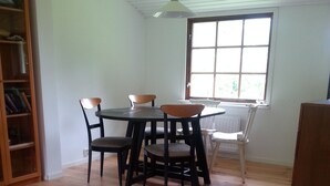 A dining area with a round table and six chairs, a wooden bookshelf, and a window with a view of greenery.