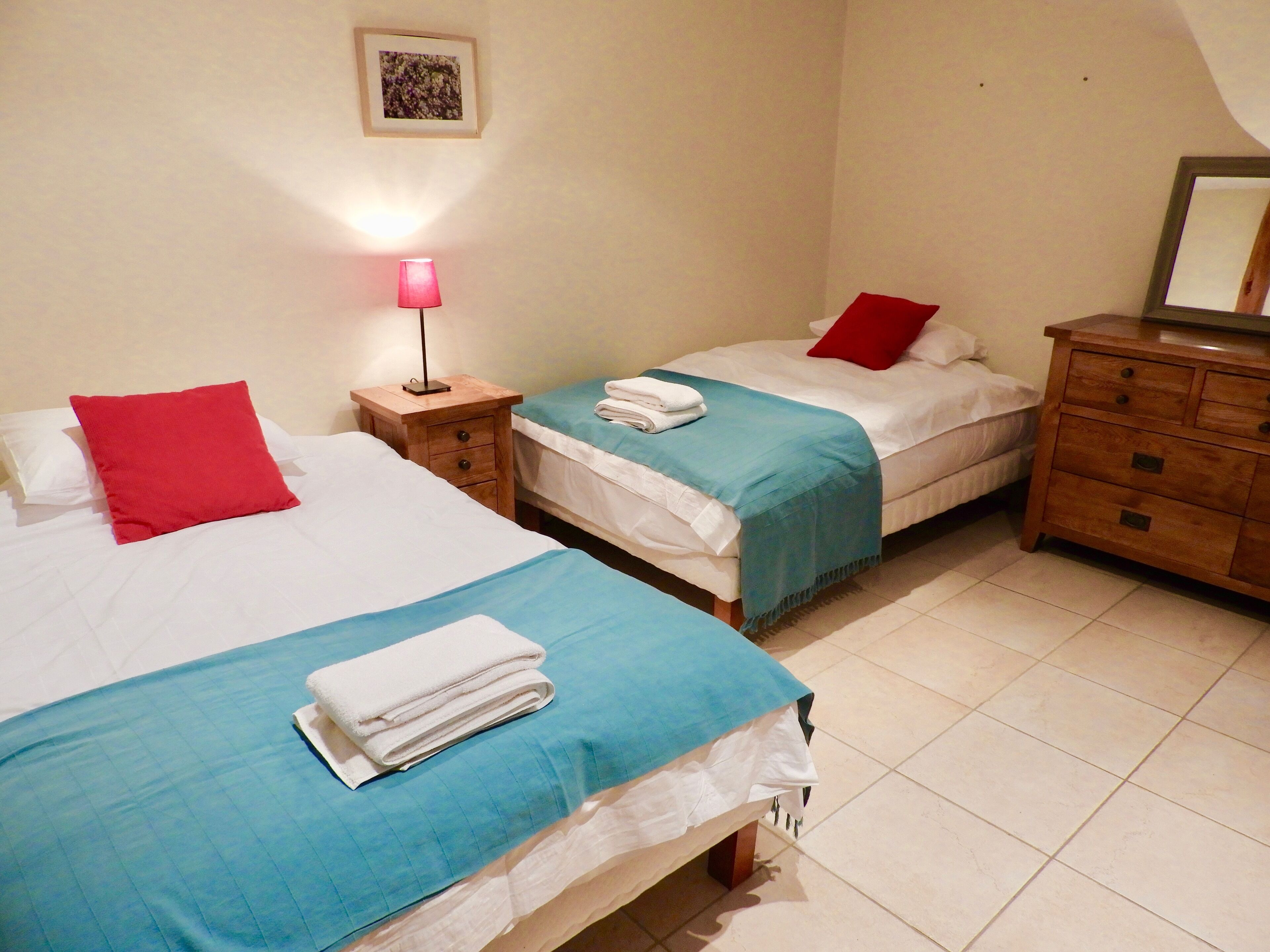 Un dormitorio con dos camas, cada una con una almohada roja, una colcha azul y una manta blanca. Entre las camas hay una mesita de noche de madera con una lámpara.