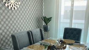 A dining area with a glass table, grey upholstered chairs, patterned wallpaper, a potted plant, and a decorative wall clock. - TIVOLI 3 BEDROOM APARTMENT, NEAR BUENAVISTA. (Barranquilla)