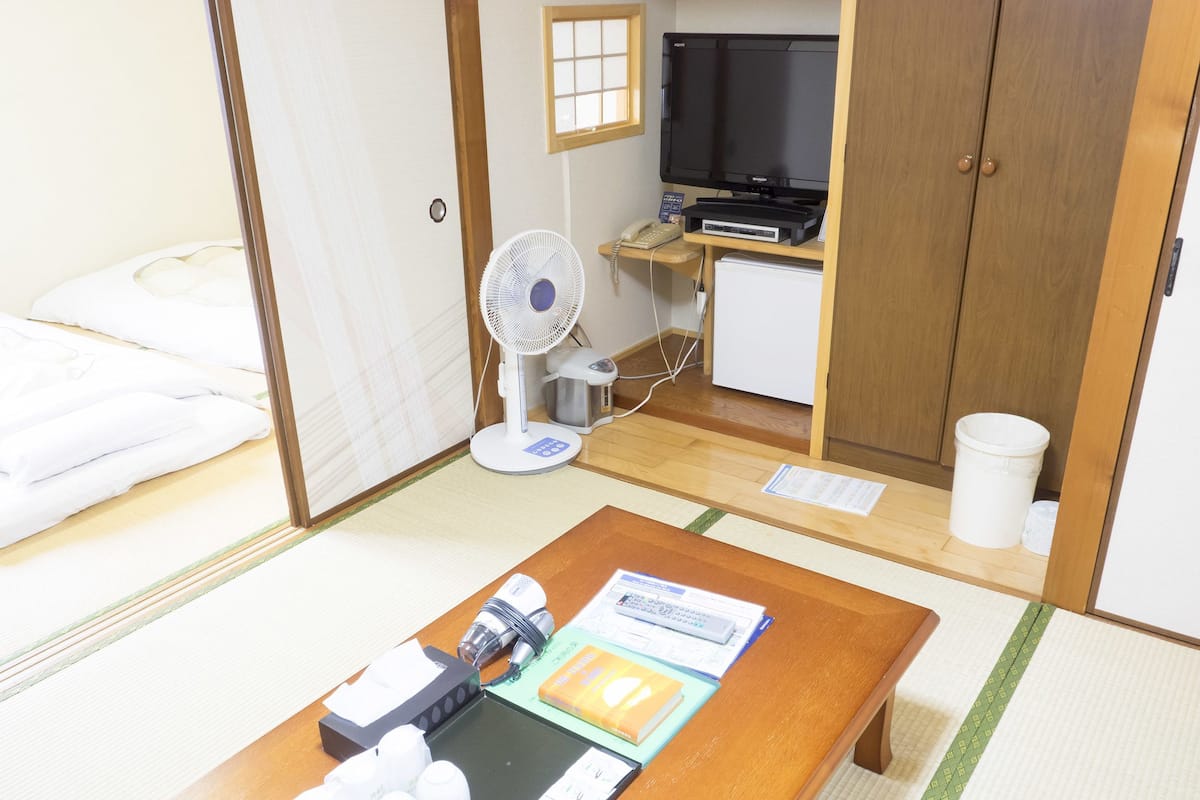 A hotel room with a bed, a wooden desk with a fan, a TV, and a small kitchenette.