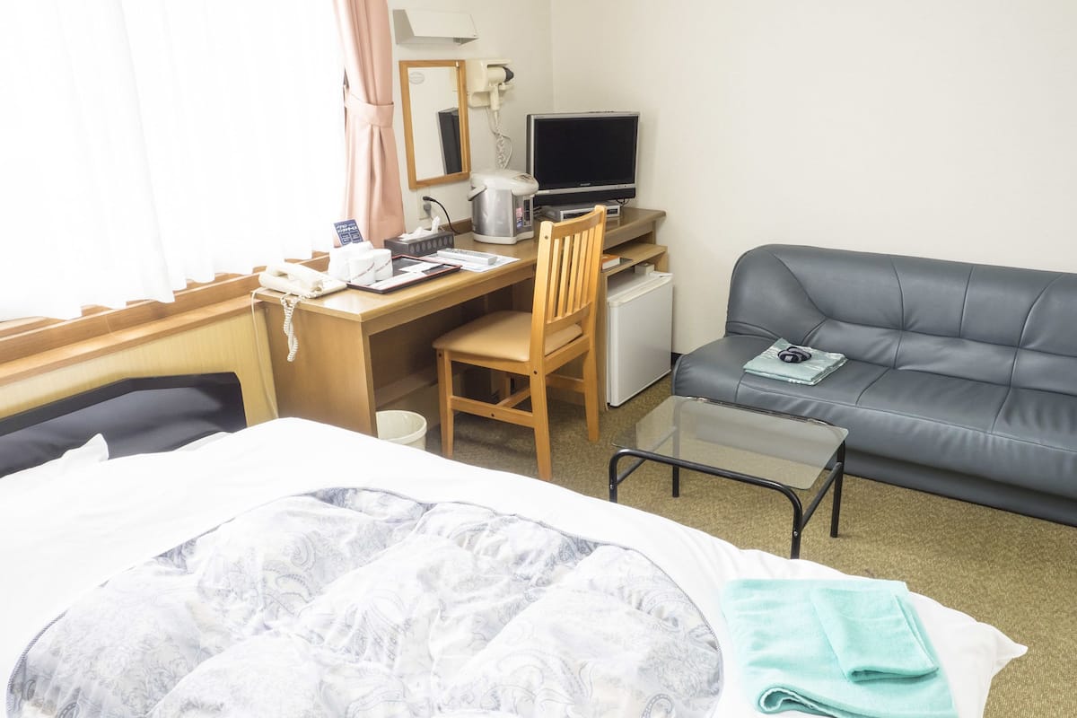 A hotel room with a bed, a desk with a computer, a chair, a grey sofa, and a small table.
