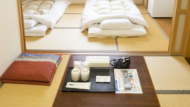 A traditional Japanese room with tatami flooring, two tatami mats, a wooden table with a tray of toiletries, and a bed with white bedding. - Hikone Station Hotel (Hikone)