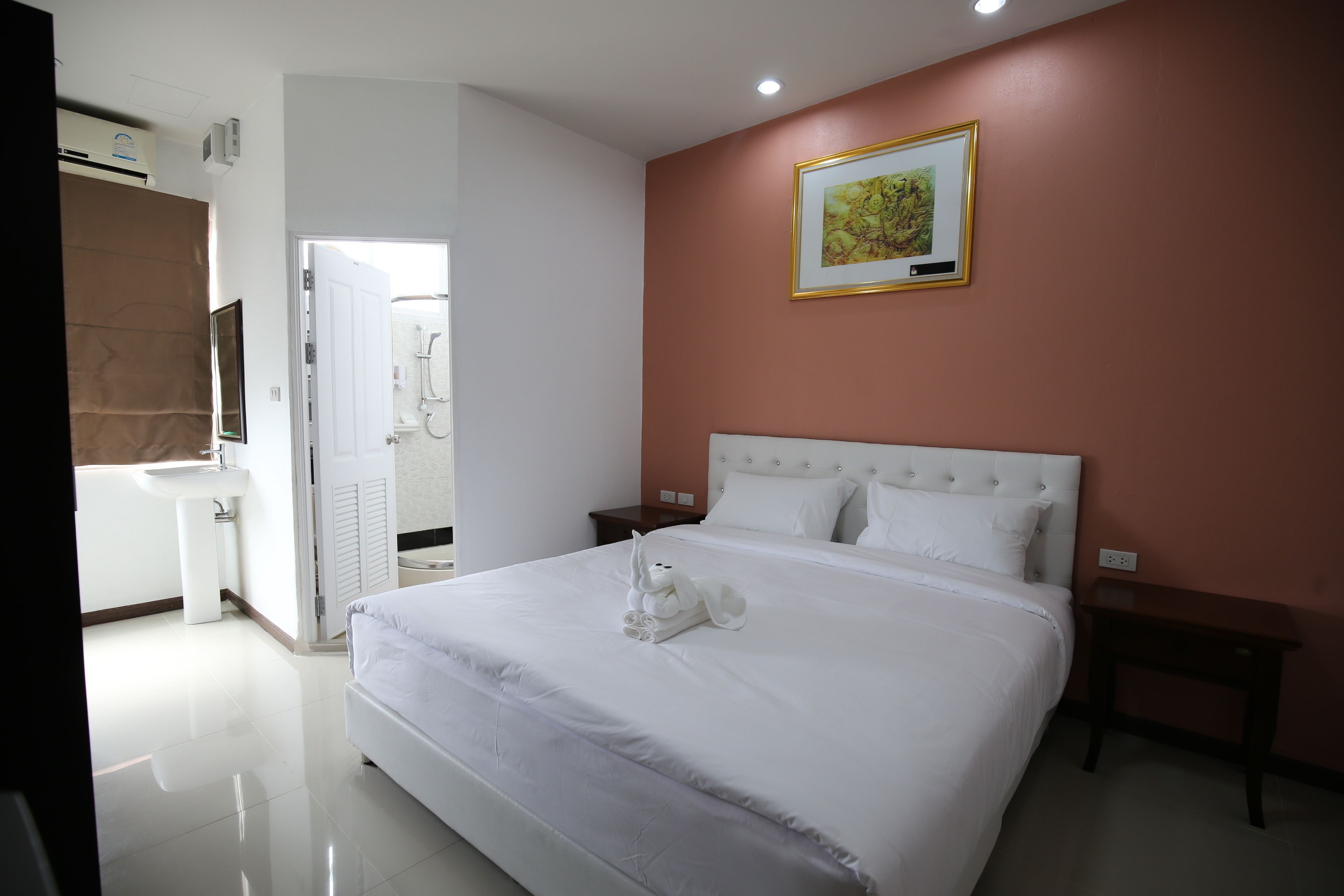 A neatly made bed with a white comforter and pillows, a bedside table, a framed picture on the wall, and a bathroom with a sink and mirror.