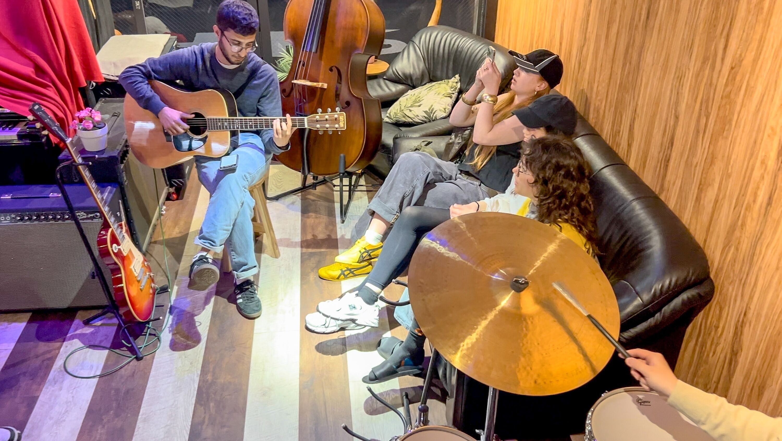 A room with a person playing an acoustic guitar, another person sitting on a couch, and a third person holding a drum.
