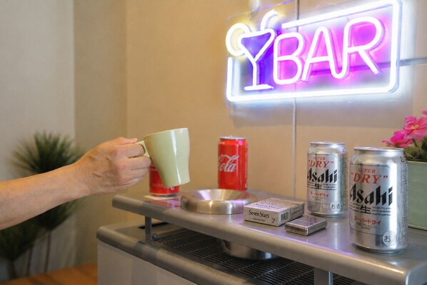 Una mano sosteniendo una taza, un letrero de neón que dice 'YBAR' y latas de cerveza Asahi sobre una barra.