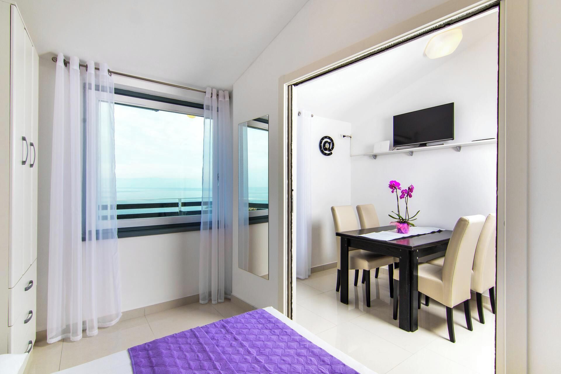 Una stanza con un letto, un tavolo da pranzo con sedie, una TV appesa al muro e vista sul mare attraverso una finestra.