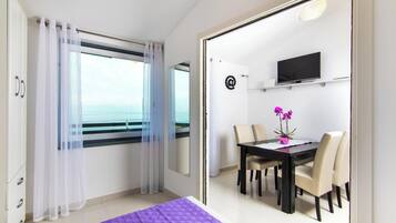 Una stanza con un letto, un tavolo da pranzo con sedie, una TV appesa al muro e vista sul mare attraverso una finestra.