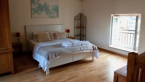 A neatly made bed with a headboard, a wooden nightstand with a lamp, a metal rack, and a window with a view of the outside. - La Forge - Apartment private terrace, overlooking the pool and Roche de Solutré (Vergisson)