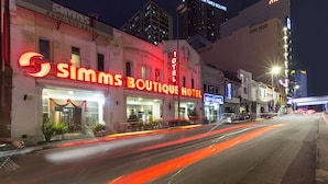 Une vue de la rue où se trouve le Simm's Boutique Hotel, avec une enseigne au néon bien visible, une entrée clairement identifiable et les bâtiments adjacents.