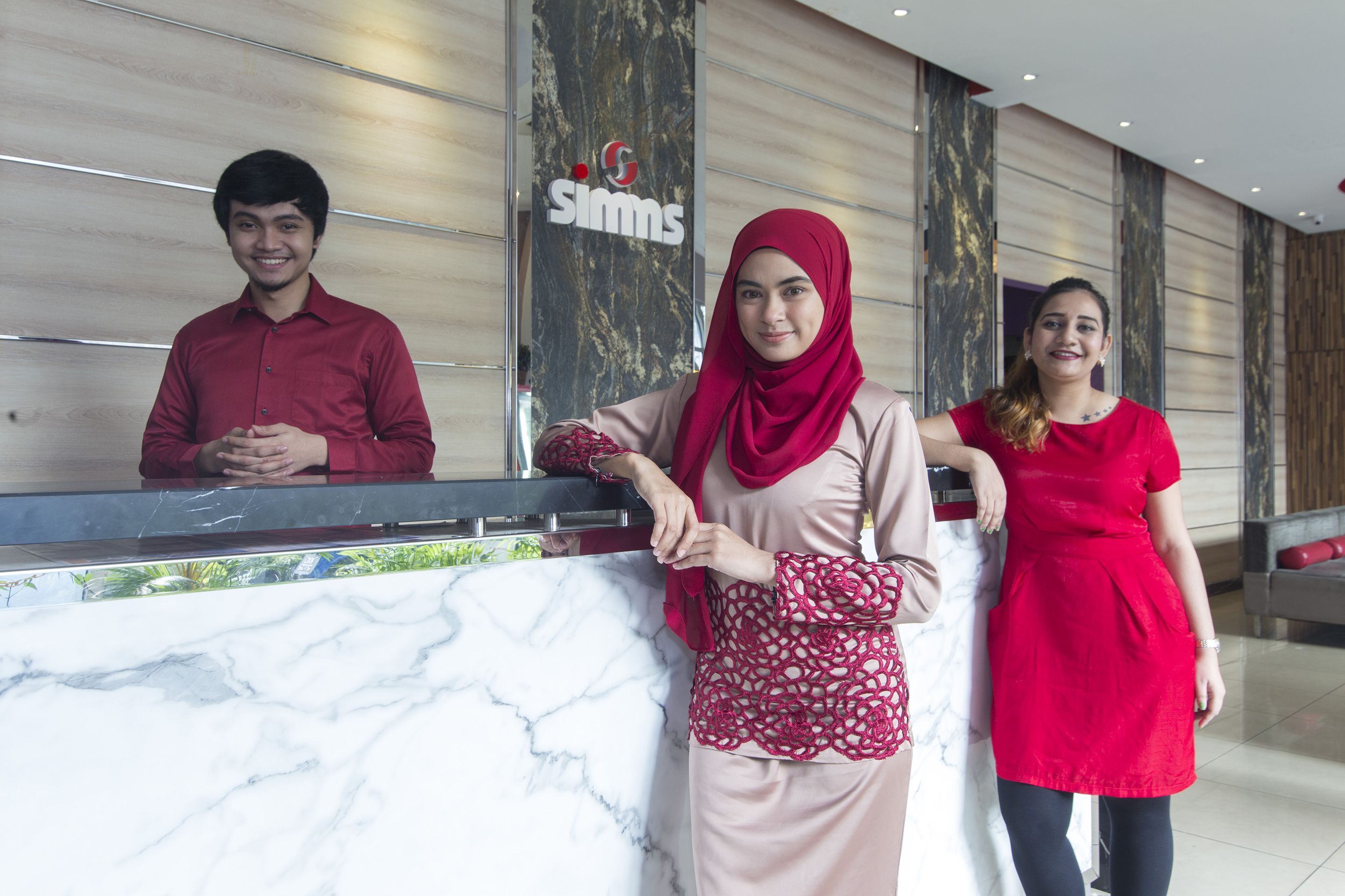 Three individuals standing at a reception desk with a marble counter and a 'SIMS' sign in the background.