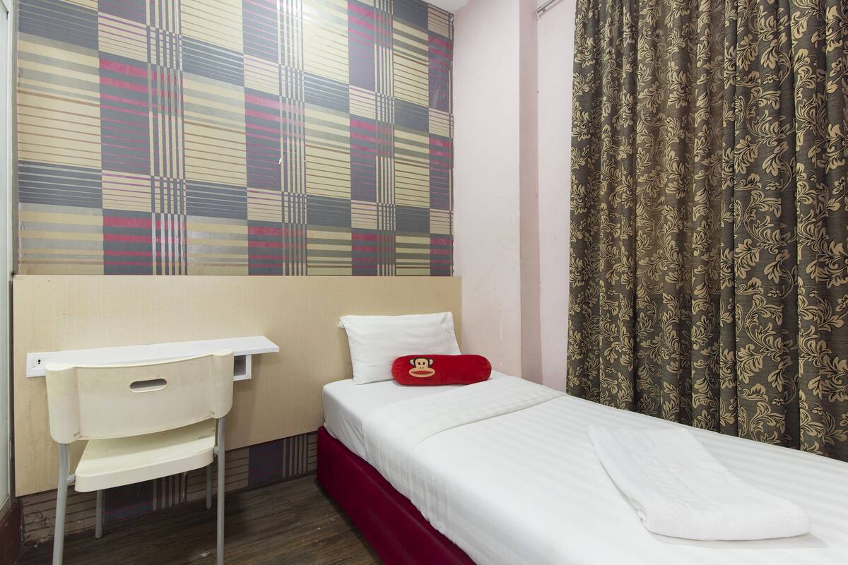 Een enkel bed met een witte matras en een rode sprei, een witte nachtkastje met een rood kussen, en een behangetje boven het bed.