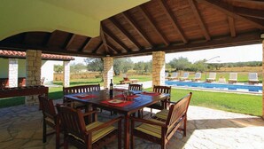 An outdoor patio with a wooden table and chairs, stone pillars, and a view of a swimming pool and lawn. - Luxury villa with heated pool and large private garden, near the sea (Peroj)