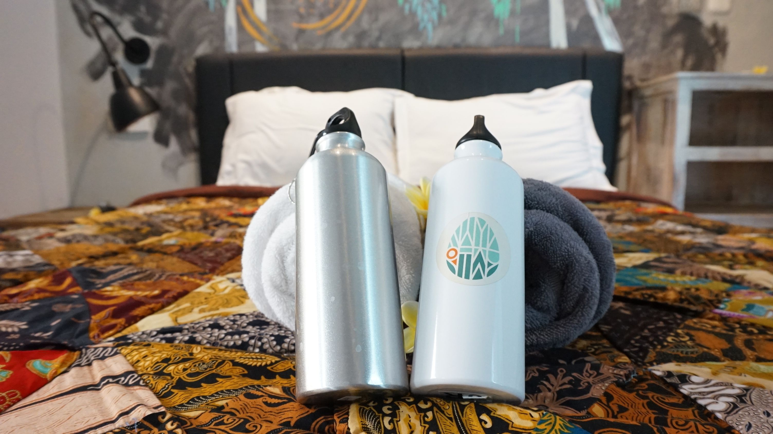 A bed with a patterned bedspread, two water bottles, and a rolled towel.