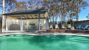 Una piscina con un porche techado, rodeada de árboles y mobiliario de exterior.