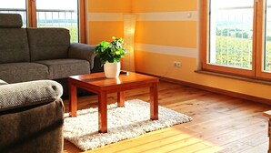 Un salón con sofá, una mesa de centro con planta, suelo de madera y ventanas amplias con vistas.