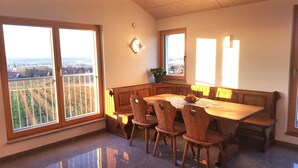 Un comedor con mesa y bancos de madera, ventanas amplias con vistas a viñedos y una planta en maceta en el alféizar.