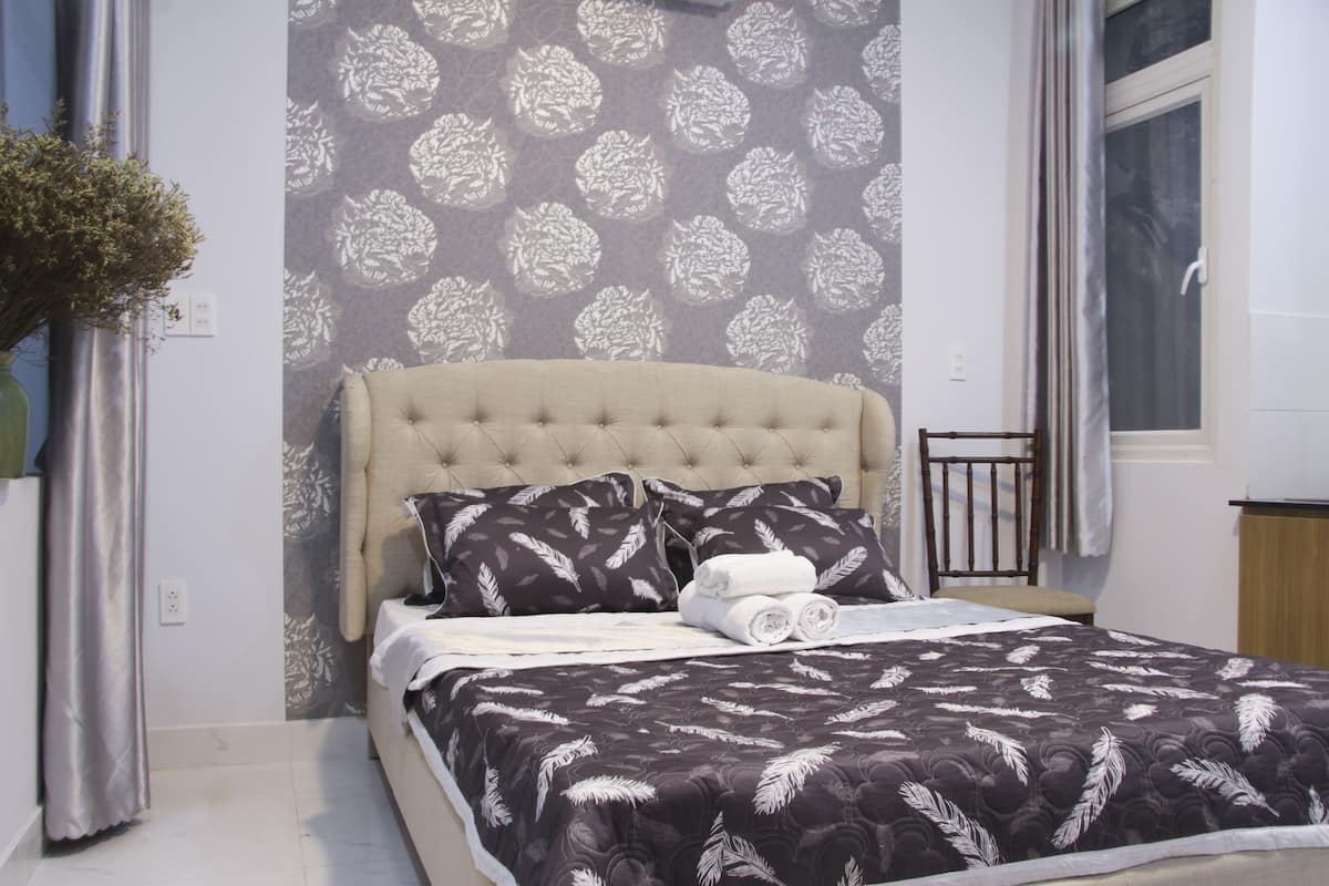 A neatly made bed with a dark patterned bedspread and matching pillows, a tufted headboard, and a wall with a floral wallpaper pattern.