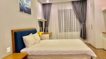 A modern hotel room with a large bed, wooden headboard, a nightstand, a bedside table, and a window with curtains.