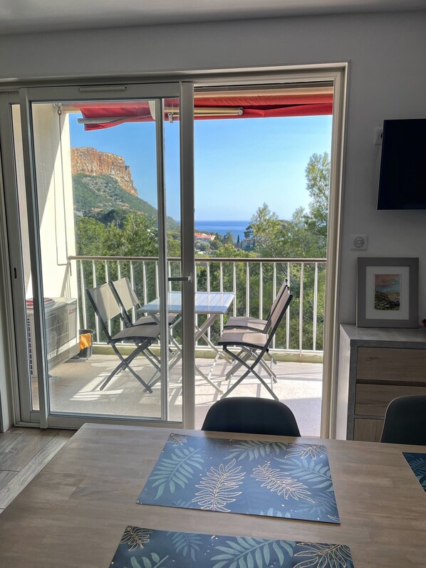 A room with a sliding glass door leading to a balcony with a table and chairs, a TV mounted on the wall, and a framed picture. - Apartment in residence, sea view and Cap Canaille (Cassis)