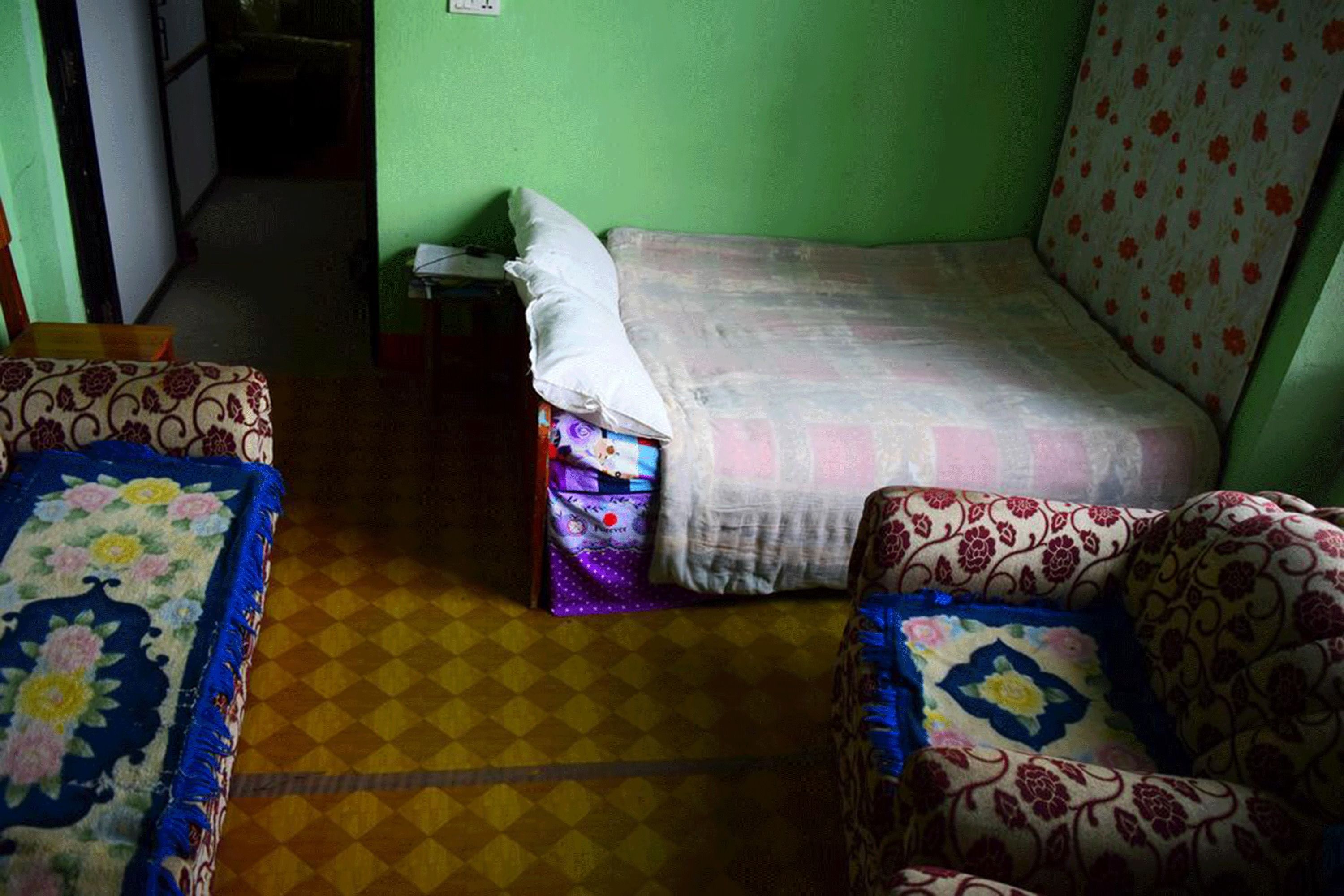Una habitación pequeña y poco iluminada, con una cama individual, una silla y una alfombra estampada.
