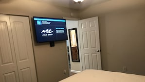 Un dormitori amb un llit, una televisió penjada a la paret que mostra un canal de música, i una porta que condueix a una altra habitació.