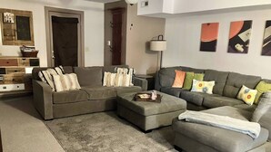 A living room with a sectional sofa, an ottoman, and a small coffee table. There is a door, a lamp, and a cabinet in the background.