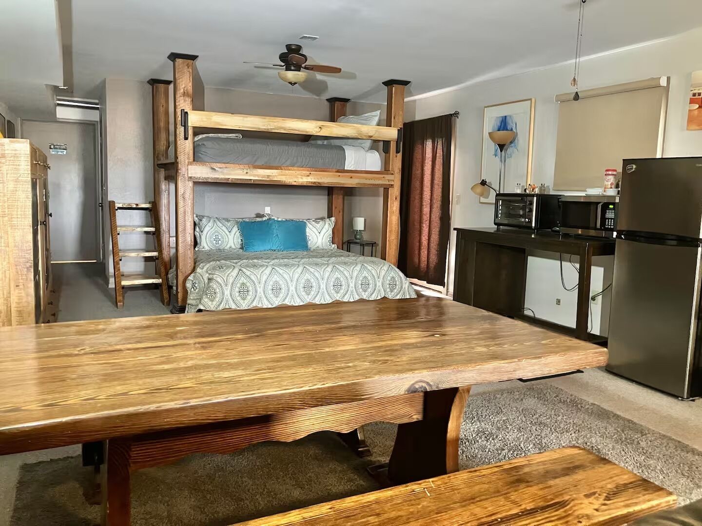 Un letto a castello con struttura in legno e biancheria a motivi, una cucina con microonde e frigorifero, e una ventola a soffitto.