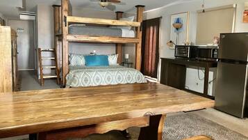Un letto a castello con struttura in legno e biancheria a motivi, una cucina con microonde e frigorifero, e una ventola a soffitto.