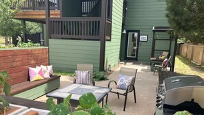 A patio area with a green building, a barbecue grill, outdoor furniture, and a garden bed with plants.